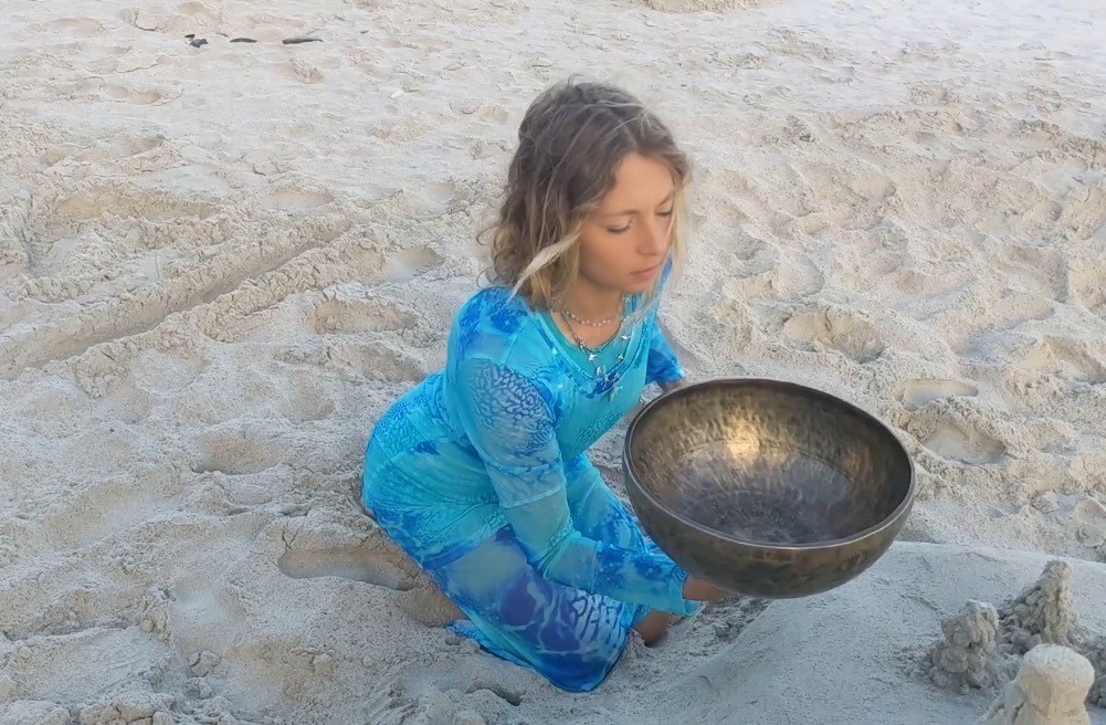 Daina Fawn in a blue dress with a singing bowl on the beach, the spirit behind Charisma